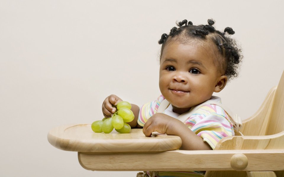 Cute Black Baby Holding Green Grapes On Wooden High Chair