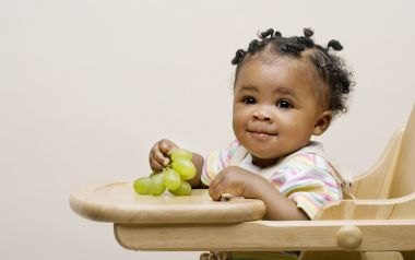Cute Black Baby Holding Green Grapes On Wooden High Chair