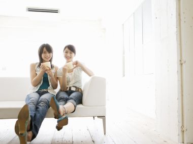 Two Girlfriends Sit On White Sofa Drinking In Bright Japanese Living Room