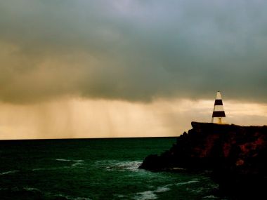 黄昏雨幕下海岸礁石上的条纹灯塔海景